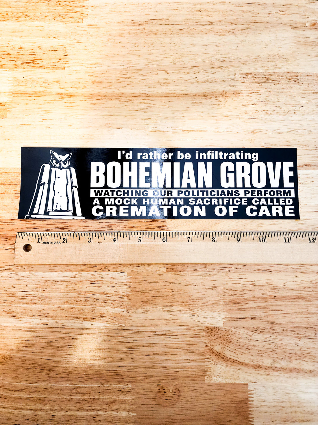 A rectangular black bumper sticker with white text and a white hooded figure graphic, placed on a wooden surface above a wooden ruler for scale.