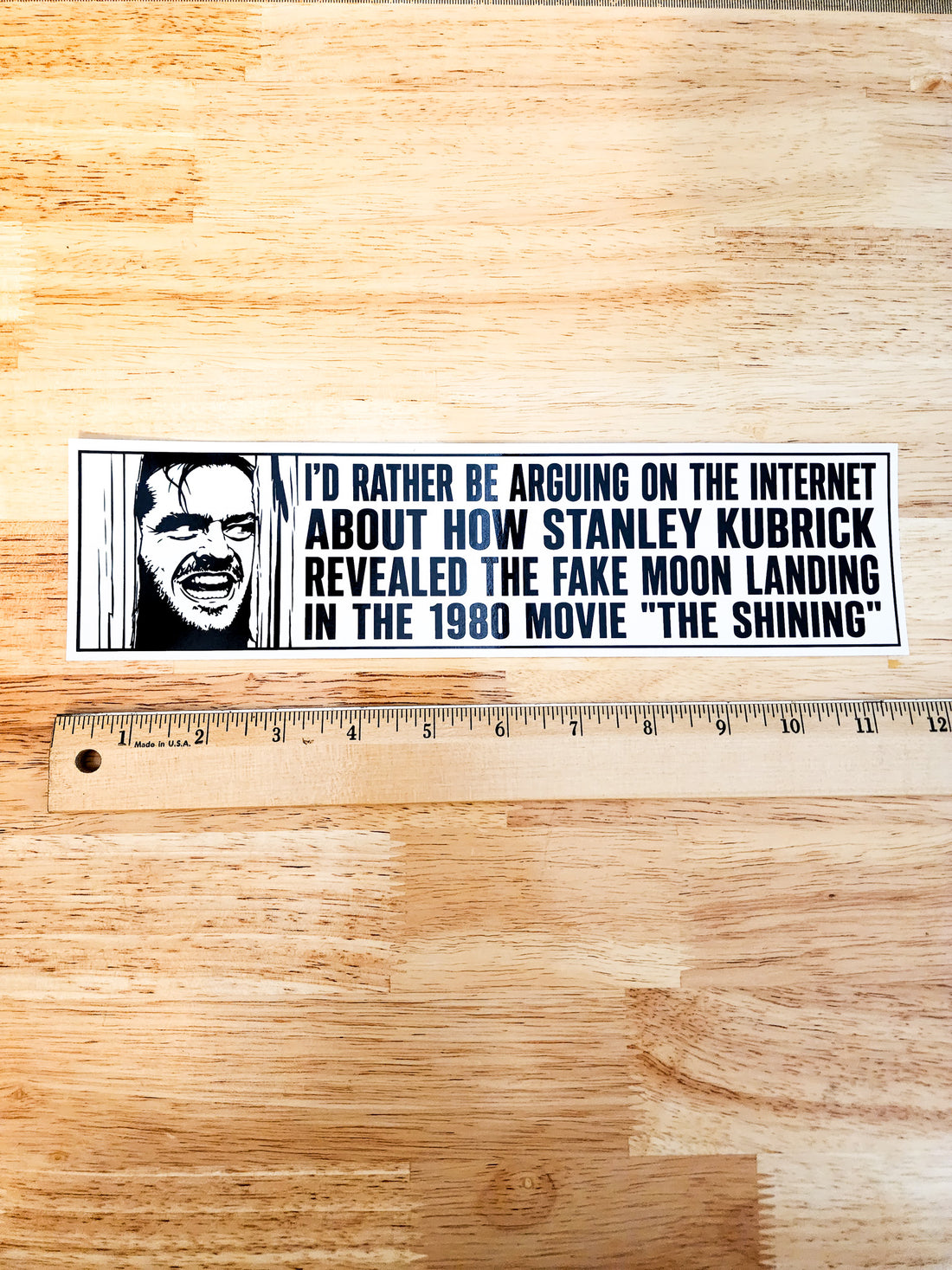 A rectangular printed sticker or decal with black text and a black-and-white illustrated face, placed on a light wood surface above a metal ruler for scale.