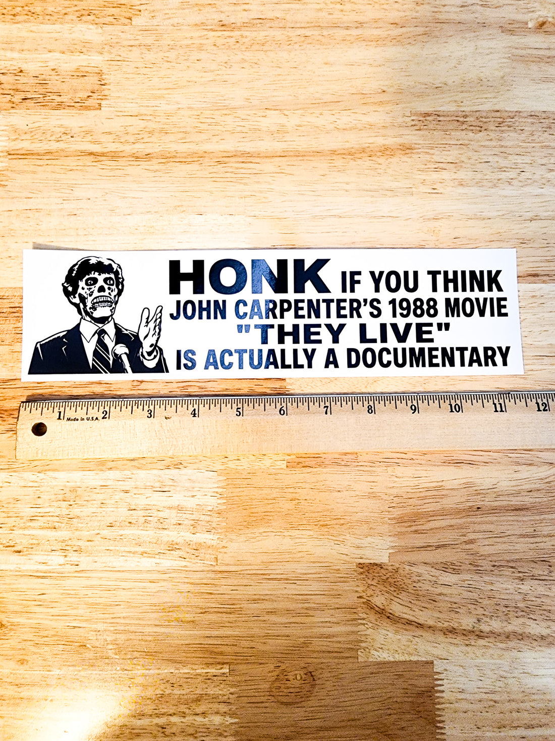 A rectangular bumper sticker with a black-and-white illustration of John Carpenter and the text &