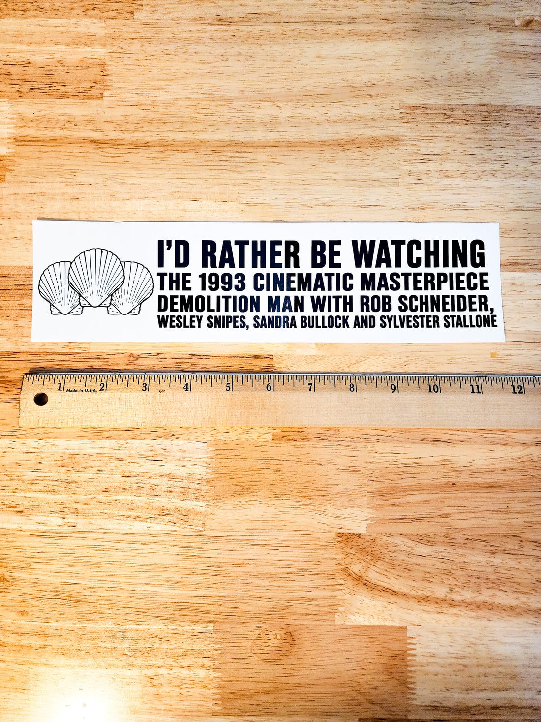 A rectangular white sticker with black text and shell illustrations, placed on a wooden surface above a wooden ruler for scale. The sticker reads: &